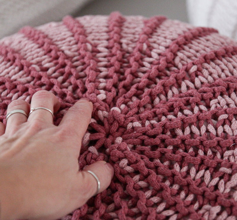 A close-up of a round, chunky-knit pouf or cushion in alternating dusty rose and light pink stripes, with a hand resting on the textured surface.