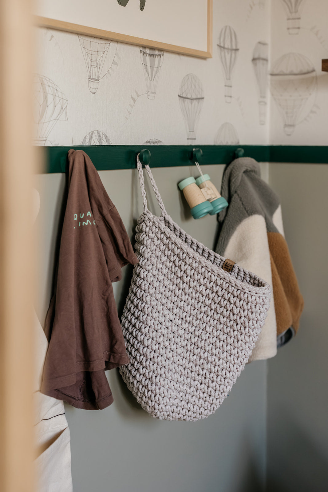 Hanging clothes including a knitted bag, hoodie, and jacket against a wall with wallpaper.