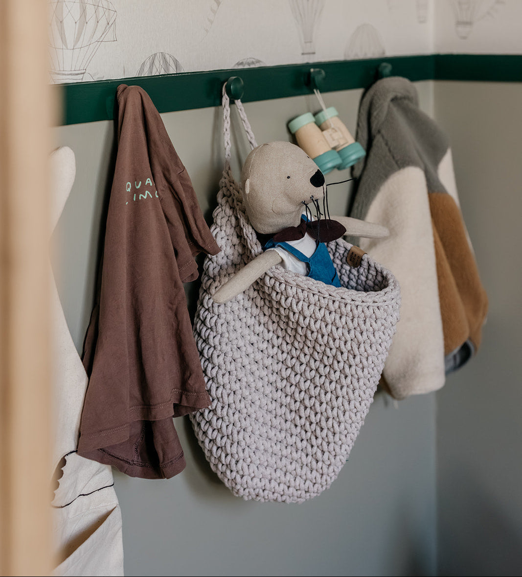 Knitted bag with toys hanging on a hook against a wall with patterned wallpaper.
