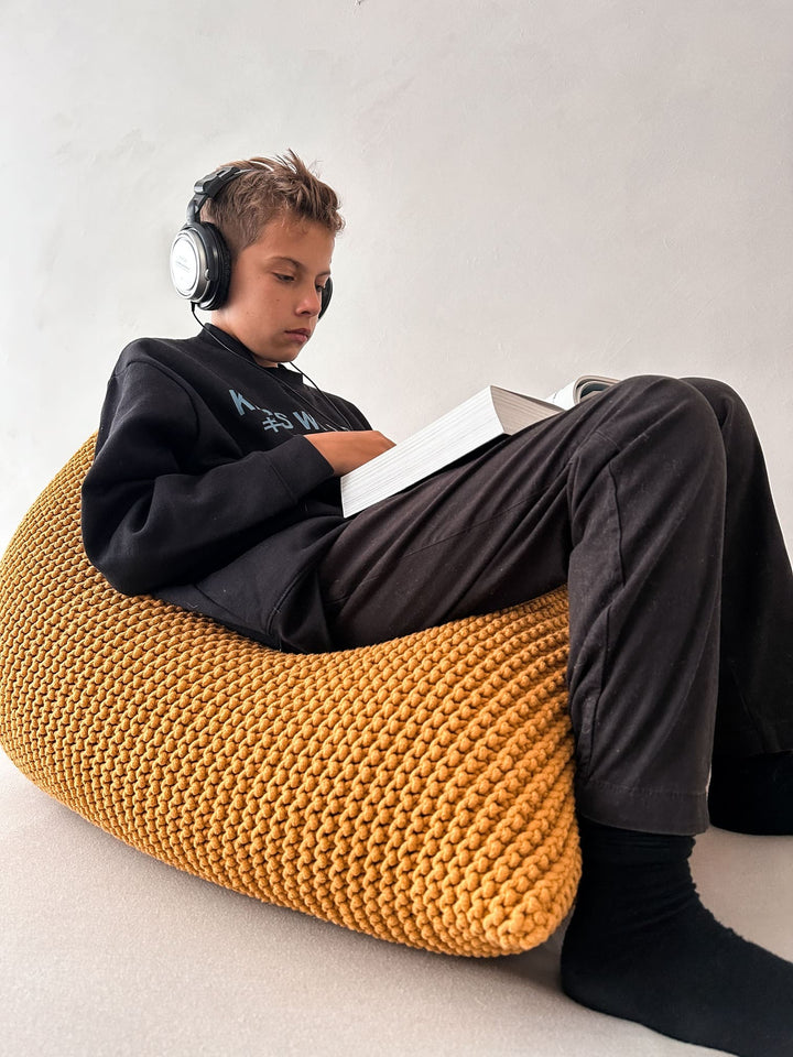 a boy relaxing on a knitted bean bag chair and listening to music, reading a book