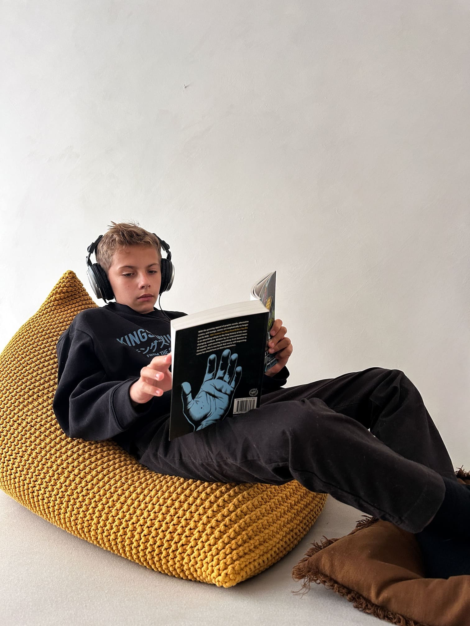a boy sitting on a yellow knitted bean bag, reading a book and listening to music