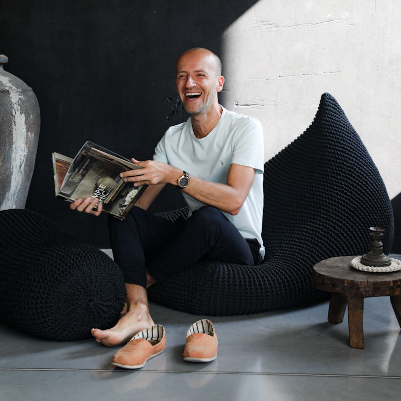Man sitting on a black knitted pouf reading a magazine in a modern interior setting.