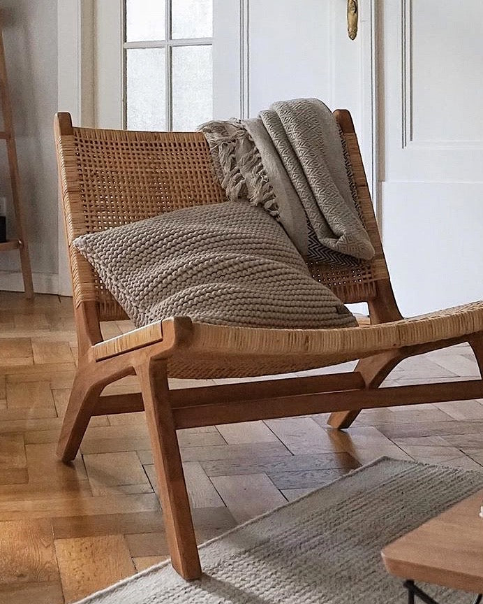 A mid-century modern wooden lounge chair with woven cane back and seat, styled with a textured beige throw blanket and matching cushion in a bright, sunlit room with parquet flooring.
