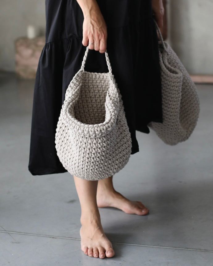 A person wearing a black dress holds a chunky crocheted shoulder bag in a neutral light gray color. The person is barefoot on a concrete floor, with a second matching bag hanging from the other hand.