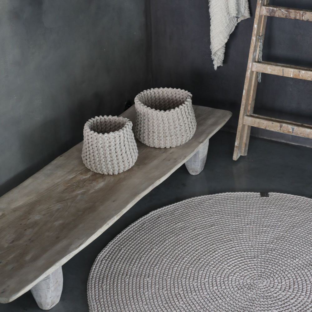 Two woven baskets on a wooden bench against a dark wall with a textured rug in front.