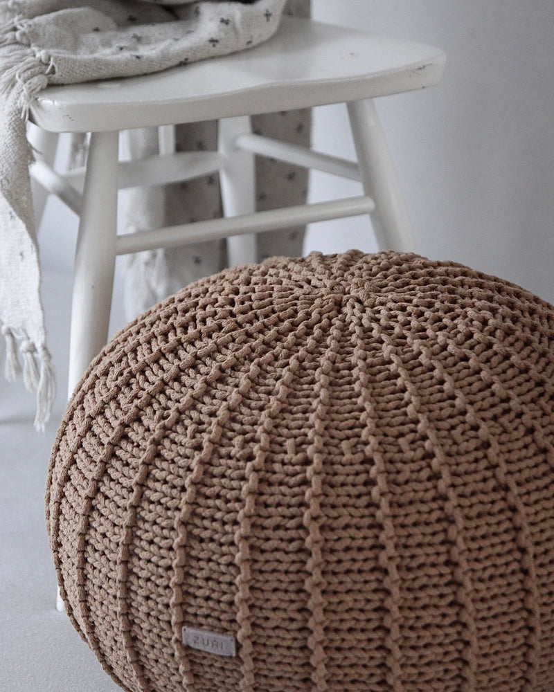 A round, chunky-knit pouf ottoman in a warm taupe-brown color, positioned in the foreground with a white wooden chair and a draped throw blanket in the background.