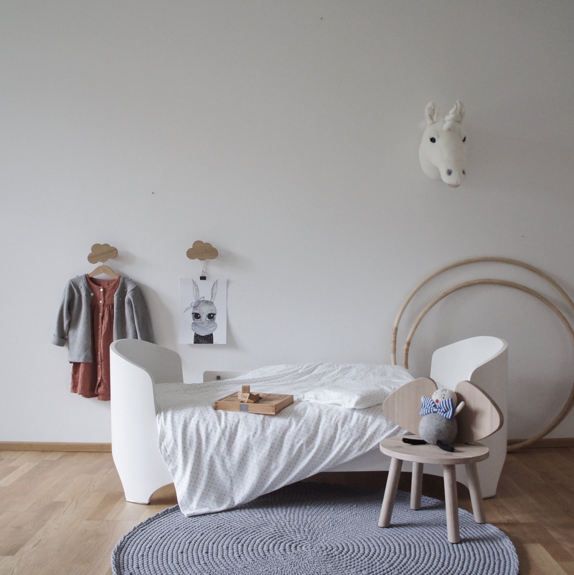 Children's room with a white bed, small table, grey crochet rug and decorative items on a light gray wall.