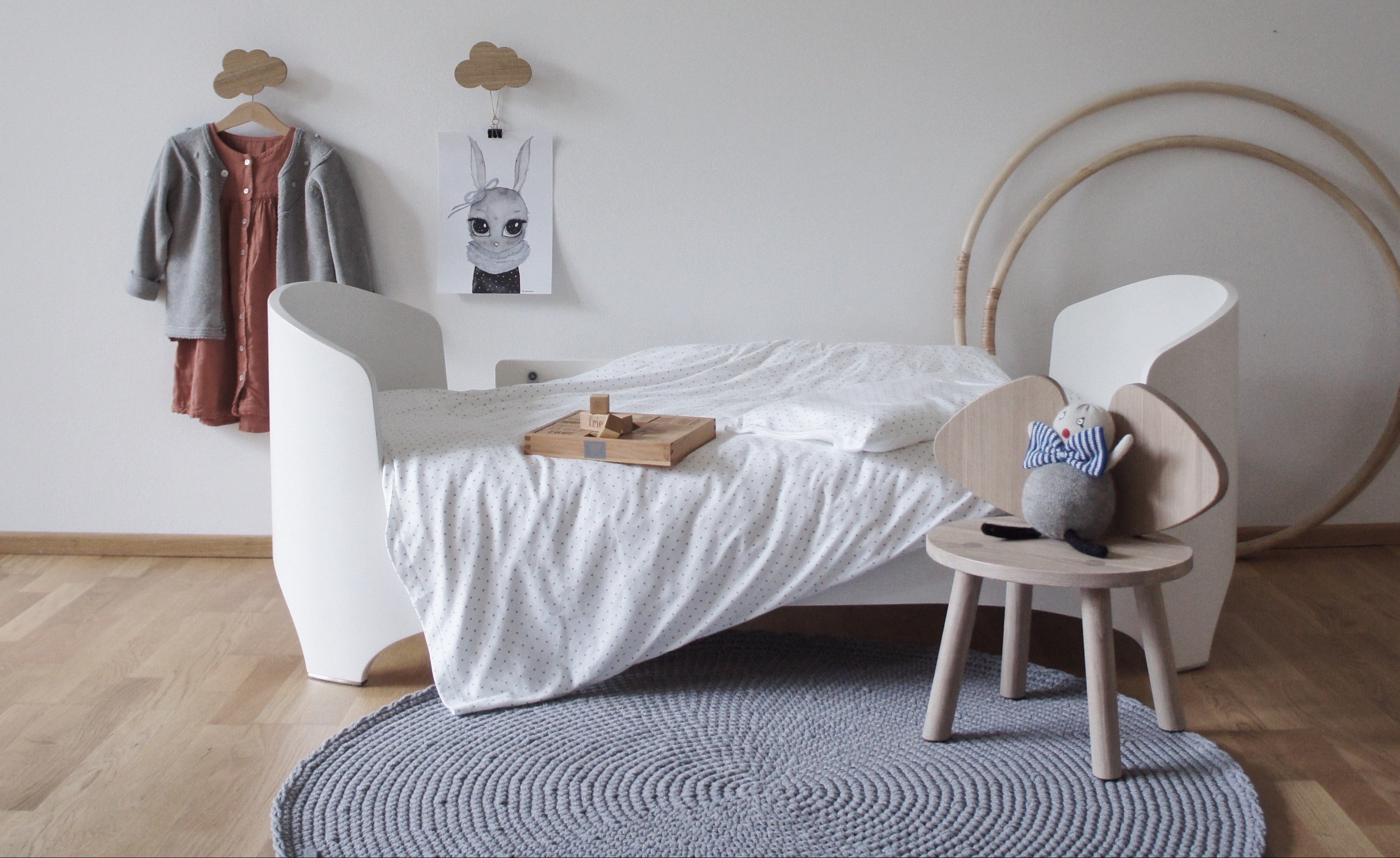 Children's room with a white bed, small table, grey crochet rug and decorative items on a light gray wall.