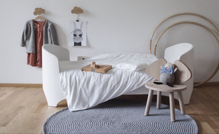 Children's room with a white bed, small table, grey crochet rug and decorative items on a light gray wall.