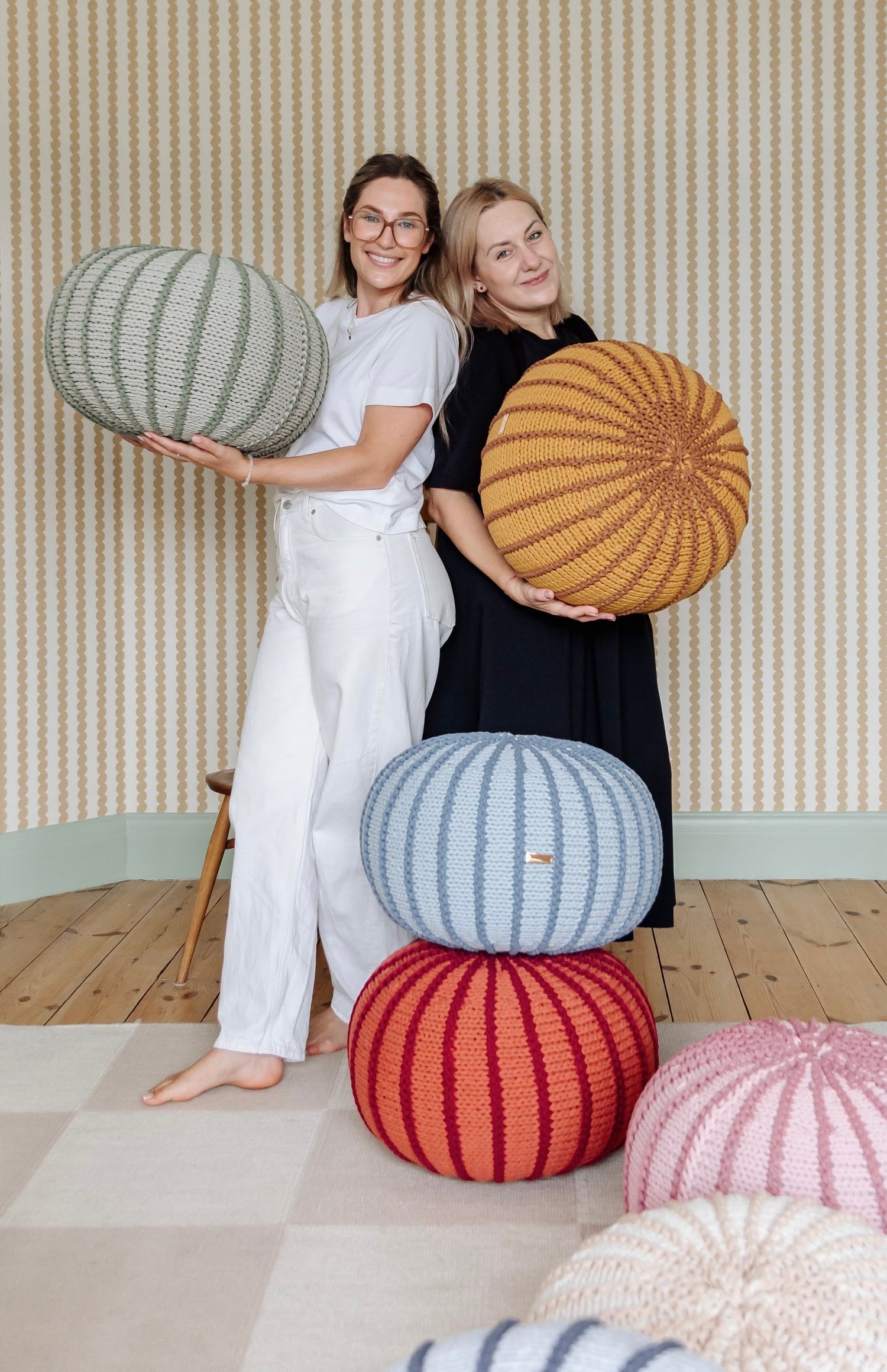 Two women standing in a room with striped wallpaper hold round knitted poufs. One pouf is light gray-green, the other is mustard yellow.